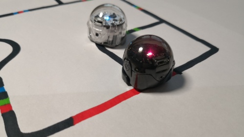 Annika Eissing / Stadtbibliothek Saarbrücken Die Szene zeigt zwei kleine, kugelförmige Roboter, die auf einem weißen Blatt positioniert sind. Der Roboter links ist transparent und der rechts ist tiefschwarz. Beide Roboter stehen auf einem weißen Hintergrund, auf dem schwarze Linien, unterbrochen von