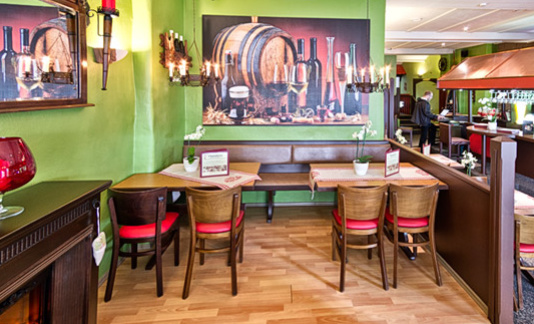 Innenraum der Flammerie mit Holztischen, grüner Wand, links ein großer Spiegel und vorne ein Bild mit einem großen Faß und verschiedenen Flaschen und Gläsern