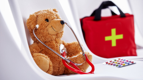Erste Hilfe Das Bild zeigt einen braunen Teddybären, der auf einem weißen Stuhl sitzt und ein rotes Stethoskop um den Hals trägt. Im Hintergrund steht eine rote Erste-Hilfe-Tasche mit einem grünen Kreuz. Auf dem Stuhl liegt neben dem Bären eine Packung bunter Pillen.
