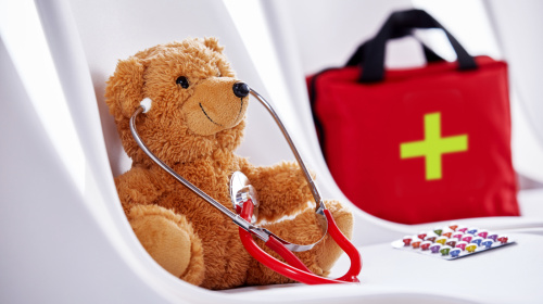 Erste Hilfe Das Bild zeigt einen braunen Teddybären, der auf einem weißen Stuhl sitzt und ein rotes Stethoskop um den Hals trägt. Im Hintergrund steht eine rote Erste-Hilfe-Tasche mit einem grünen Kreuz. Auf dem Stuhl liegt neben dem Bären eine Packung bunter Pillen.