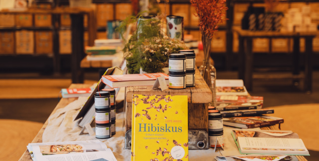 Im Vordergrund ist ein Tisch mit Rezeptbüchern. Im Hintergrund sind große, hölzerne Wandregale, die zum Teil mit Kisten und zum Teil mit Gewürzdosen gefüllt sind.
